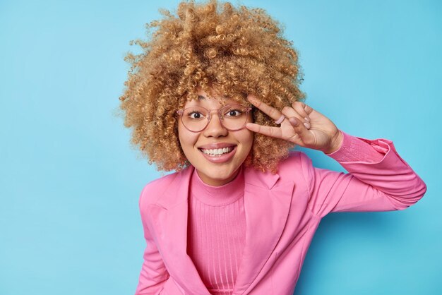 La bella donna d'affari allegra con i capelli ricci fa un gesto di pace sui sorrisi degli occhi indossa ampiamente gli occhiali giacca formale rosa posa su sfondo blu rimane sempre positiva e felice