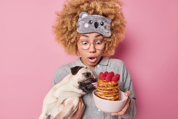 La bella donna dai capelli ricci scioccata guarda sorprendente mentre il suo cane cerca di mangiare frittelle vestite in pigiama e maschera per dormire posa con l'animale domestico preferito su sfondo rosa L'ora della colazione a casa