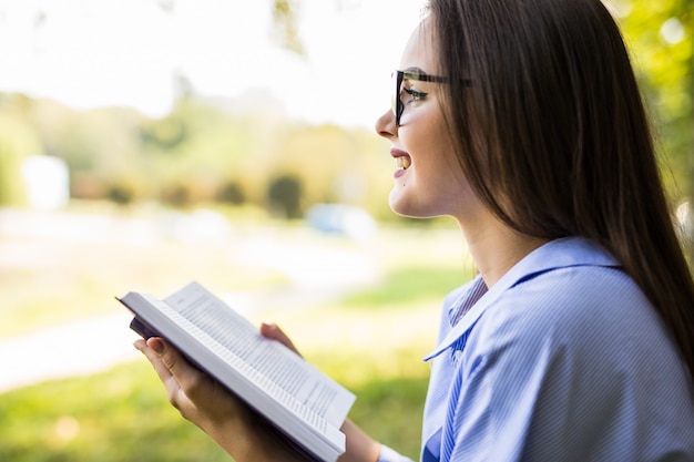 La bella donna dai capelli scuri con gli occhiali legge il libro contro il parco verde estivo.