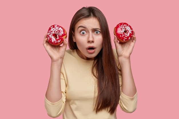 La bella donna dai capelli scuri imbarazzata tiene le ciambelle, ha un'espressione spaventata, si sente perplessa, sta su sfondo rosa