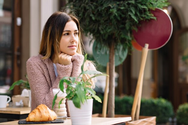 La bella donna è seduta in mensa vestita con un maglione caldo e aspetta qualcuno. Sta guardando da parte. Si riscalda con una bevanda calda. Giornata autunnale, ritratto all'esterno, incontro.