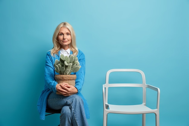 La bella donna europea rugosa sicura di sé calma sente la nostalgia che la malinconia tiene con le pose di cactus in vaso con la sedia vuota