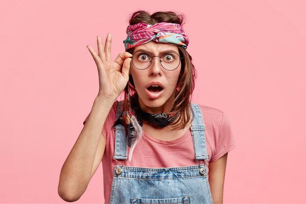 La bella donna hippy sorpresa e sorpresa guarda attraverso gli occhiali, non può credere alle cattive notizie, vestita in tuta e fascia per capelli, isolata sul muro rosa dello studio