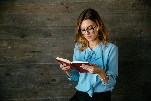 La bella donna intelligente bella in occhiali che legge il libro interessante, sembra pensierosa