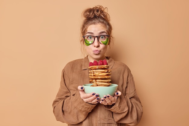 La bella donna meravigliata tiene le labbra piegate applica cerotti di idrogel verde sotto gli occhi per ridurre il gonfiore vestita con indumenti da notte tiene deliziose frittelle con sciroppo ha una gustosa colazione applica dischetti di bellezza