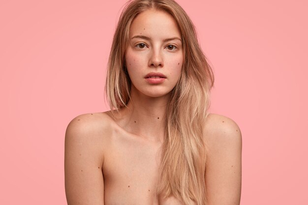 La bella donna mezza nuda mostra un corpo snello e perfetto, ha lunghi capelli chiari, guarda con espressione seria direttamente alla telecamera, isolata sul muro rosa
