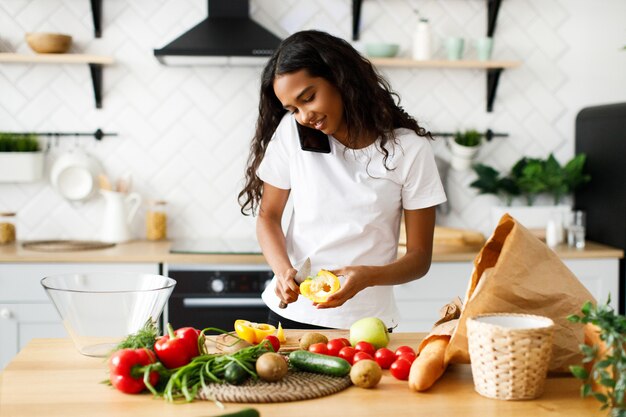 La bella donna mulatta sta cucinando un pasto dalle verdure fresche sulla cucina moderna e sta parlando al telefono