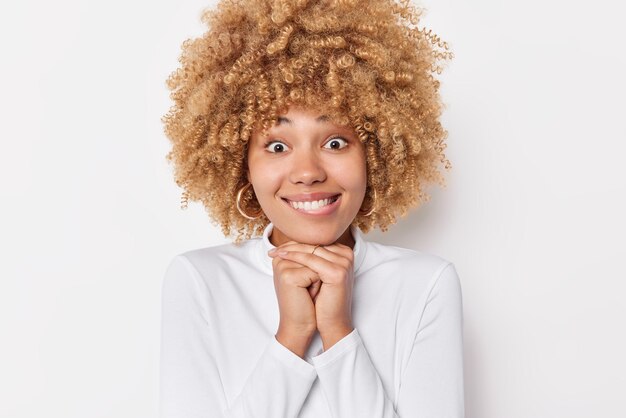 La bella donna ottimista tiene le mani sotto il mento sorride a trentadue denti ha un'espressione curiosa del viso indossa pose casual a collo alto al coperto su sfondo bianco. Concetto di emozioni e sentimenti felici