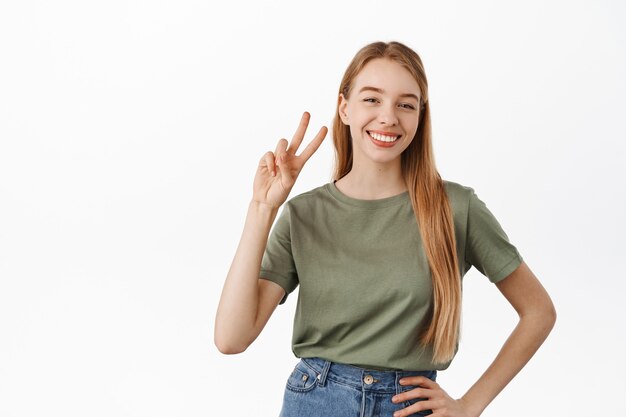 La bella donna positiva mostra il segno v di pace e sorride felice con i denti bianchi perfetti, in piedi in maglietta e jeans contro il muro dello studio