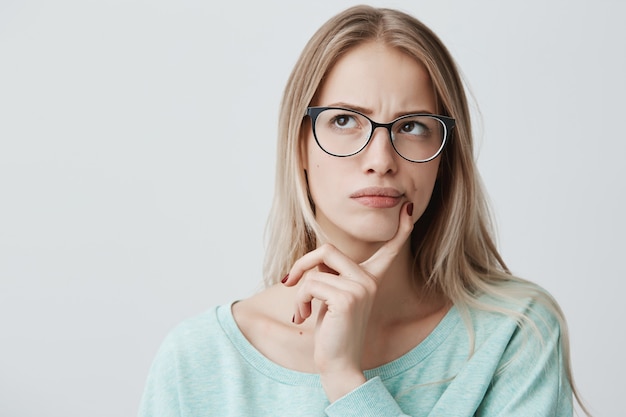 la bella donna premurosa ha lunghi capelli biondi con occhiali eleganti, guarda da parte con espressione pensierosa, pianifica qualcosa nei prossimi fine settimana, posa contro il muro bianco. Femmina perplessa