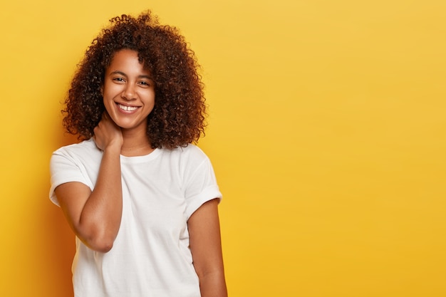 La bella donna riccia tocca il collo, sorride con gioia, ha un aspetto civettuolo, gode del tempo libero, indossa una maglietta bianca casual, parla casualmente con qualcuno, esprime emozioni positive isolate sul muro giallo