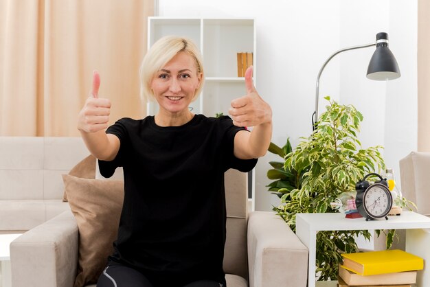 La bella donna russa bionda sorridente si siede sui pollici in su della poltrona con due mani