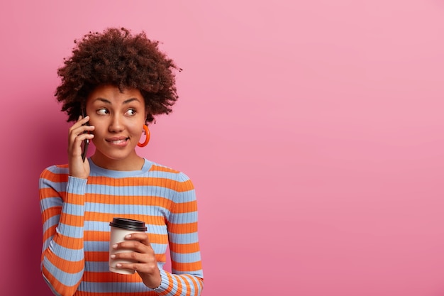 La bella donna seria con i capelli afro ha una conversazione telefonica, guarda da parte, si morde le labbra, tiene lo smartphone vicino all'orecchio