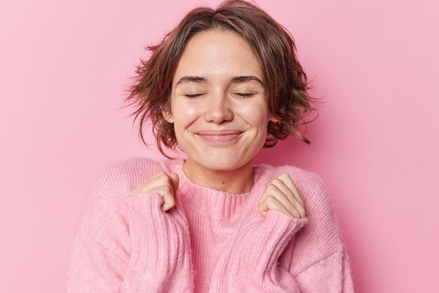 La bella donna soddisfatta scuote le mani tiene gli occhi chiusi attende sorrisi a sorpresa indossa con gioia un maglione morbido isolato su sfondo rosa. La femmina castana allegra anticipa per smth piacevole