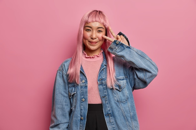 La bella donna sorridente augura fortuna e pace, mostra il segno della vittoria, fa un viso carino e seducente, si diverte, capelli rosa dritti tinti lunghi, indossa una giacca di jeans