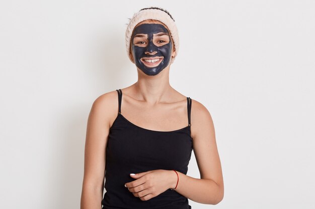 La bella donna sorridente con la maschera facciale dell'argilla nera sul fronte che sta contro la parete bianca con il sorriso affascinante, ragazza sveglia che fa le procedure cosmetiche a casa, sembra felice.