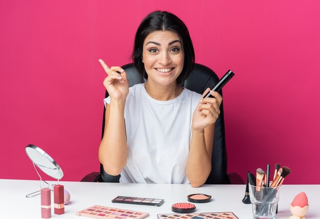 La bella donna sorridente si siede al tavolo con gli strumenti per il trucco che tengono i punti del pennello in polvere verso l'alto