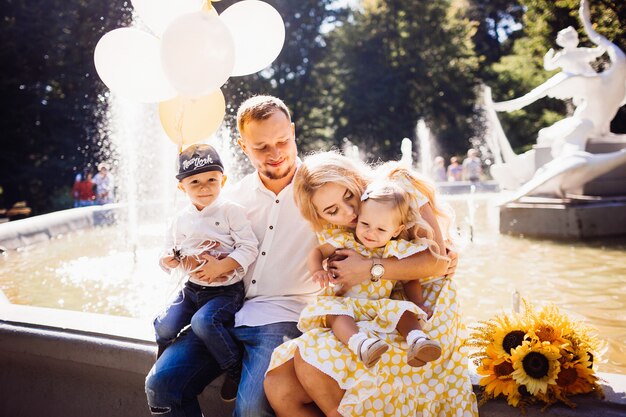 La bella famiglia vestita con gli stessi vestiti si siede sulla fontana con i loro bambini e palloncini gialli