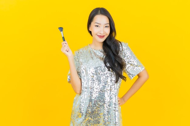 La bella giovane donna asiatica del ritratto con compone il cosmetico della spazzola su giallo