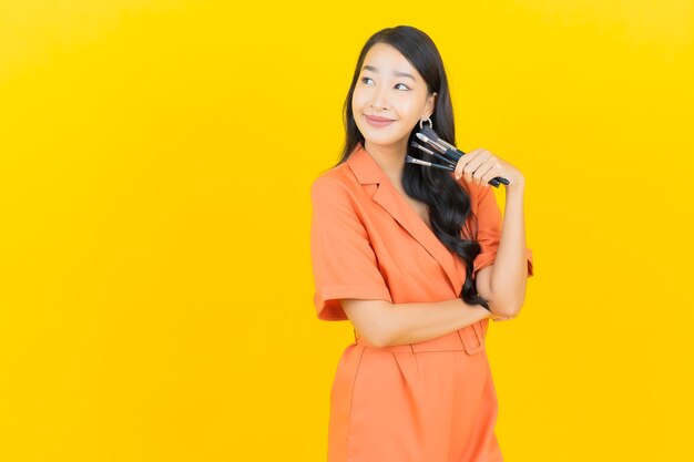 La bella giovane donna asiatica del ritratto con cosmetico compone la spazzola su colore giallo