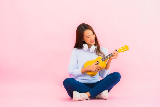 La bella giovane donna asiatica del ritratto gioca l'ukulele sulla parete isolata di colore rosa