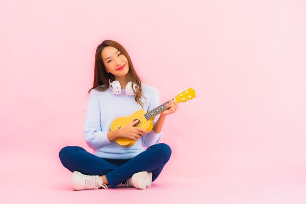 La bella giovane donna asiatica del ritratto gioca l'ukulele sulla parete isolata di colore rosa