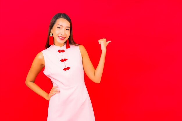 La bella giovane donna asiatica del ritratto indossa i vestiti cinesi del capodanno sulla parete rossa