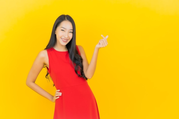 La bella giovane donna asiatica del ritratto indossa il sorriso rosso del vestito con l'azione sulla parete gialla