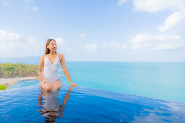 La bella giovane donna asiatica del ritratto si rilassa il tempo libero di sorriso intorno alla piscina all'aperto con l'oceano del mare nella vacanza di viaggio