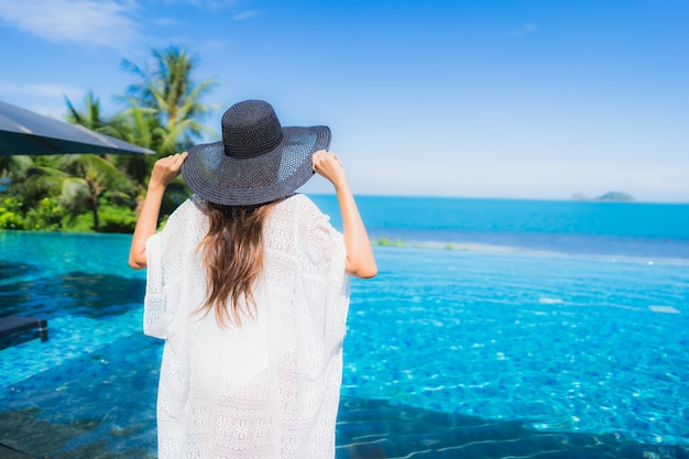 La bella giovane donna asiatica del ritratto si rilassa nella piscina all'aperto di lusso nell'oceano quasi del mare della spiaggia della località di soggiorno dell'hotel