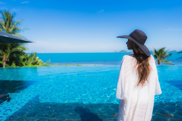 La bella giovane donna asiatica del ritratto si rilassa nella piscina all'aperto di lusso nell'oceano quasi del mare della spiaggia della località di soggiorno dell'hotel