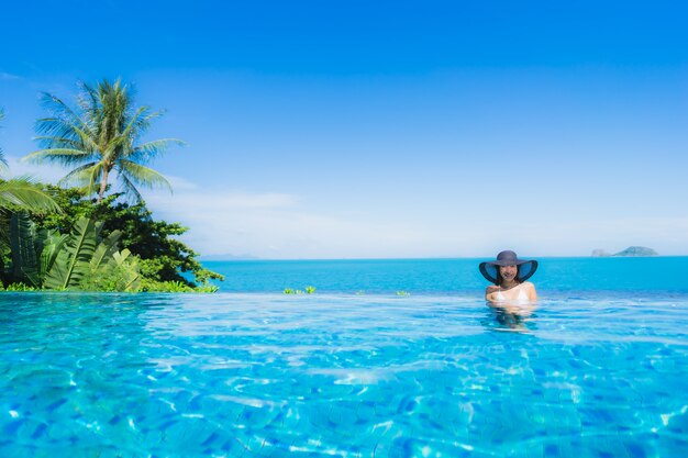 La bella giovane donna asiatica del ritratto si rilassa nella piscina all'aperto di lusso nell'oceano quasi del mare della spiaggia della località di soggiorno dell'hotel