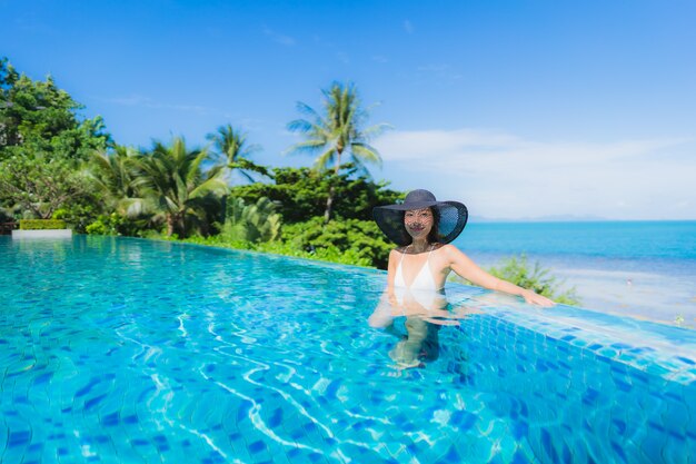 La bella giovane donna asiatica del ritratto si rilassa nella piscina all'aperto di lusso nell'oceano quasi del mare della spiaggia della località di soggiorno dell'hotel