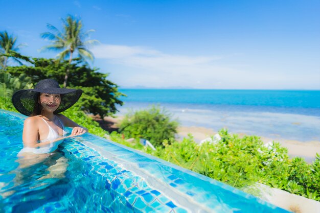 La bella giovane donna asiatica del ritratto si rilassa nella piscina all'aperto di lusso nell'oceano quasi del mare della spiaggia della località di soggiorno dell'hotel
