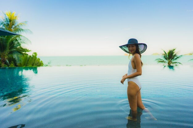 La bella giovane donna asiatica del ritratto si rilassa nella piscina all'aperto di lusso nell'oceano quasi del mare della spiaggia della località di soggiorno dell'hotel