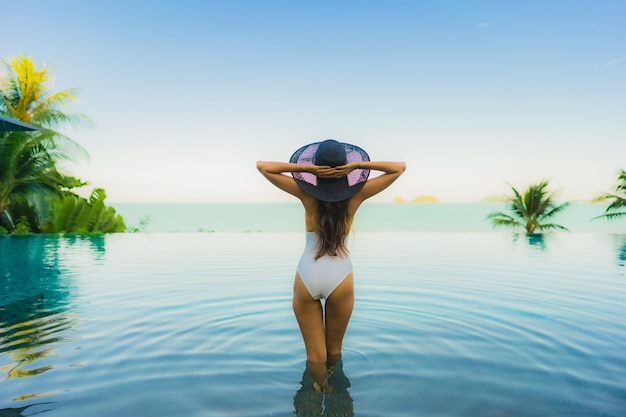 La bella giovane donna asiatica del ritratto si rilassa nella piscina all'aperto di lusso nell'oceano quasi del mare della spiaggia della località di soggiorno dell'hotel