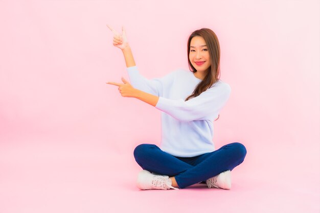 La bella giovane donna asiatica del ritratto si siede sul pavimento con la parete isolata di colore rosa