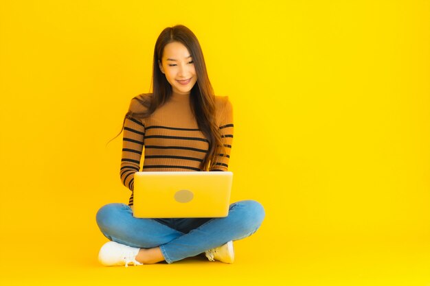 La bella giovane donna asiatica del ritratto si siede sul pavimento per il computer portatile o il computer di uso sulla parete gialla