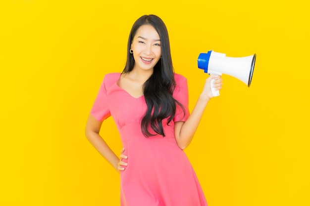 La bella giovane donna asiatica del ritratto sorride con il megafono sulla parete gialla