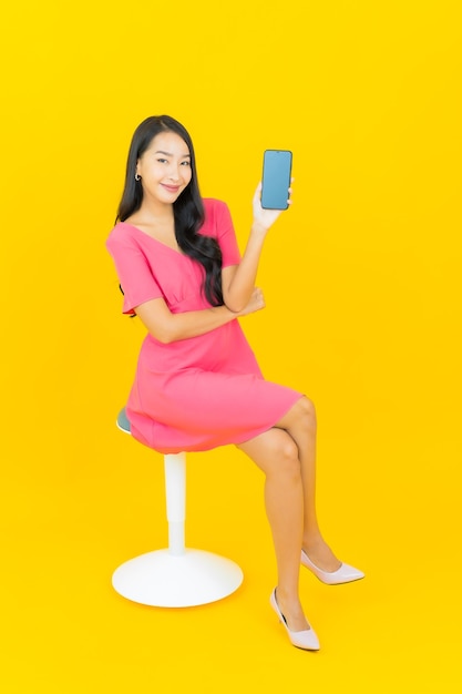 La bella giovane donna asiatica del ritratto sorride con il telefono cellulare astuto sulla parete gialla