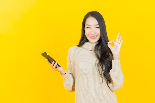La bella giovane donna asiatica del ritratto sorride con il telefono cellulare astuto sulla parete gialla