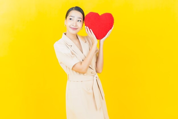 La bella giovane donna asiatica del ritratto sorride con la forma del cuscino del cuore sulla parete di colore