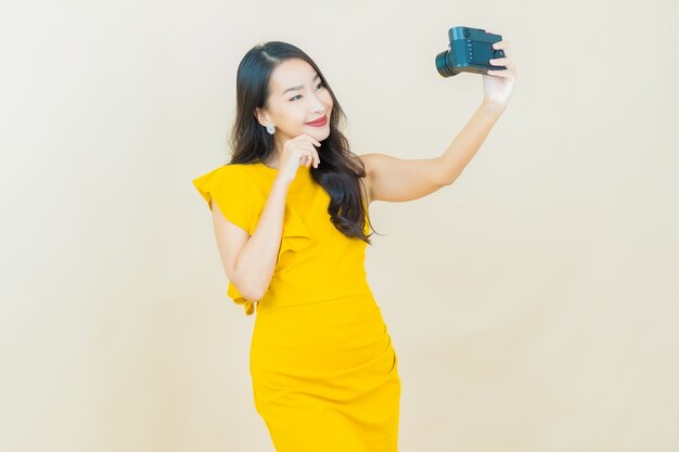 La bella giovane donna asiatica del ritratto usa la macchina fotografica sulla parete beige