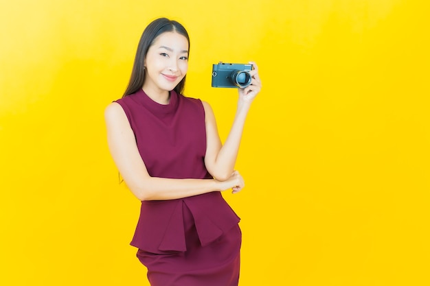 La bella giovane donna asiatica del ritratto usa la macchina fotografica sulla parete gialla