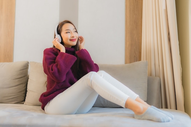 La bella giovane donna asiatica del ritratto usa le cuffie per ascoltare musica sul divano nell'interno del soggiorno