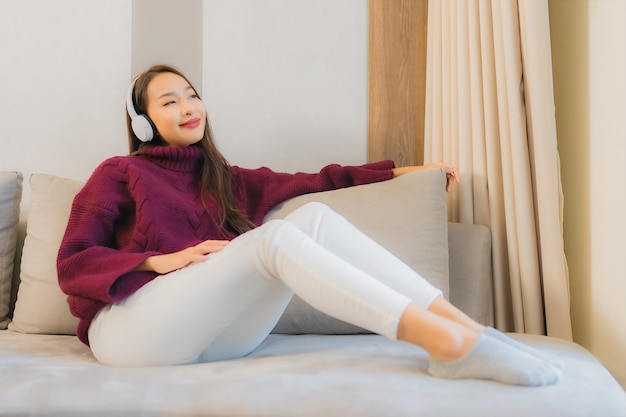 La bella giovane donna asiatica del ritratto usa le cuffie per ascoltare musica sul divano nell'interno del soggiorno