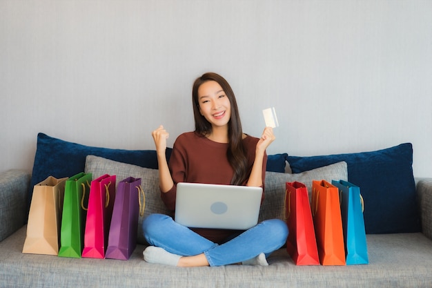 La bella giovane donna asiatica del ritratto utilizza il computer portatile e la carta di credito per lo shopping online sul divano nell'interno del soggiorno