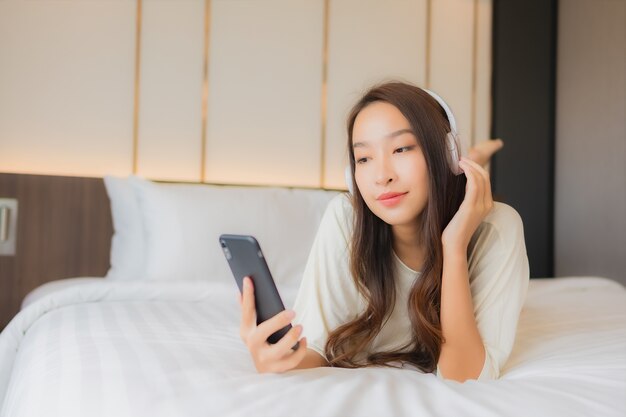 La bella giovane donna asiatica del ritratto utilizza il telefono cellulare astuto con la cuffia per ascoltare la musica in camera da letto