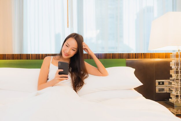 La bella giovane donna asiatica del ritratto utilizza il telefono cellulare astuto sul letto nell'interno della camera da letto