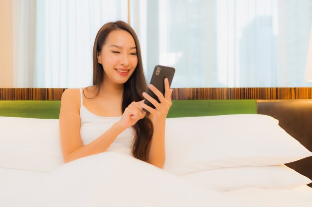 La bella giovane donna asiatica del ritratto utilizza il telefono cellulare astuto sul letto nell'interno della camera da letto